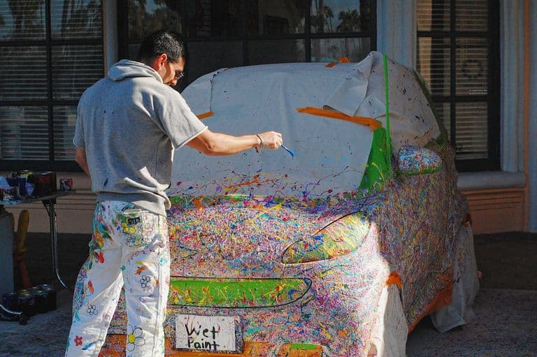 A man splatter-paints a car covered in protective sheets, creating a vibrant, abstract design across the vehicle's surface