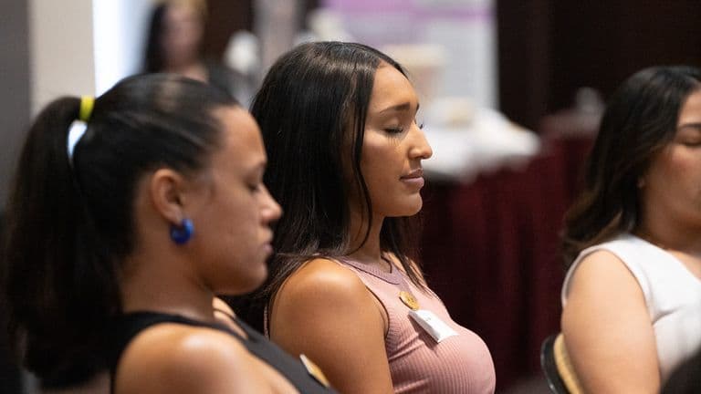 Three women sit with eyes closed in quiet meditation during a group session.