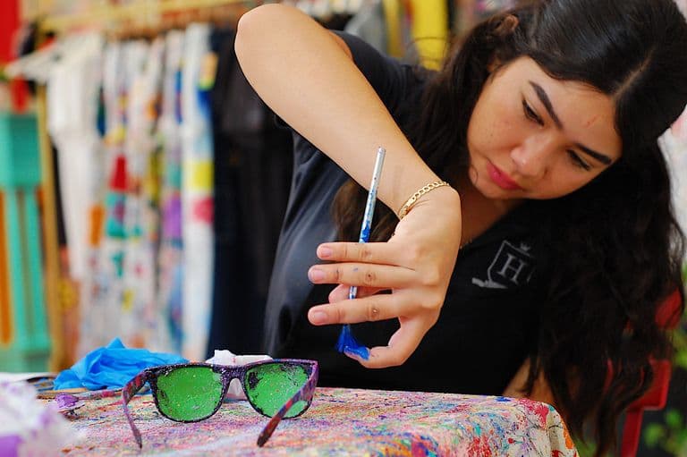 A woman carefully paints a pair of green-lensed sunglasses with a small brush, surrounded by colorful splatters on a table.