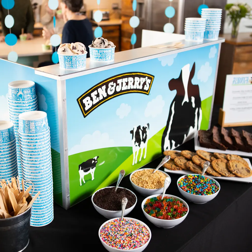 A Ben & Jerry's ice cream station with scoops in cups, an array of toppings in bowls, and stacks of branded cups. People are enjoying ice cream in the background.