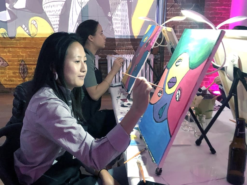 Two women painting abstract art at easels, each deeply focused on their work in a neon-lit room with drinks on the table.