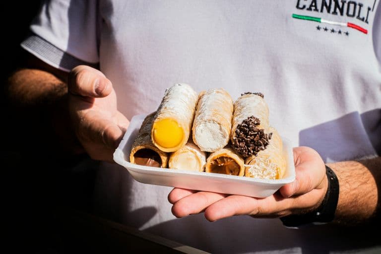 Person holding a tray of colorful cannoli with assorted fillings and toppings, dusted with powdered sugar.