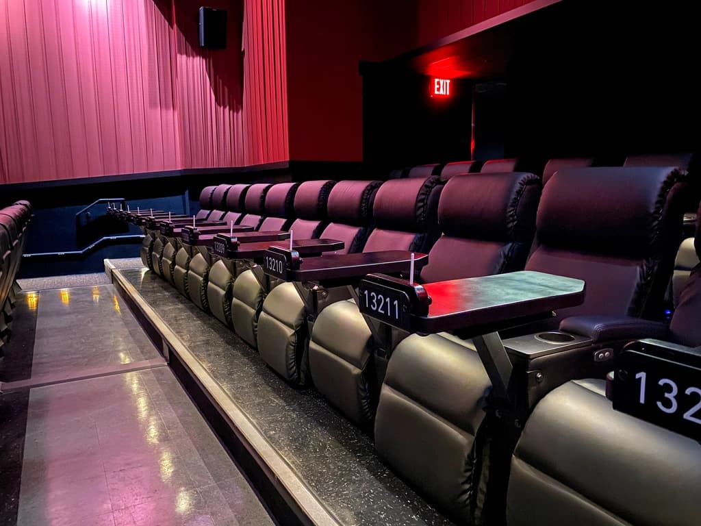 Rows of plush reclining theater seats with attached trays, each labeled with a seat number, creating a luxurious cinema atmosphere.
