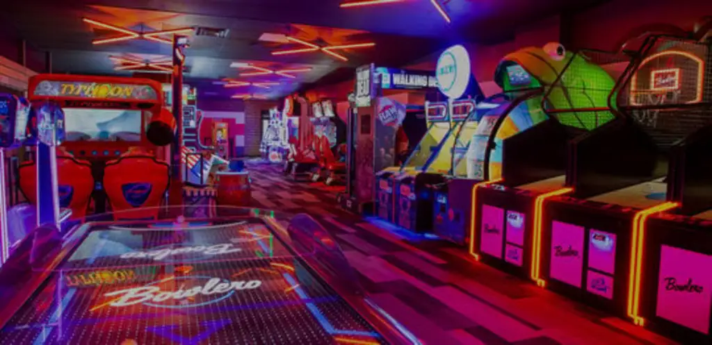 A lively arcade filled with various games, including racing simulators, basketball hoops, and air hockey, all brightly lit with neon and LED lights.
