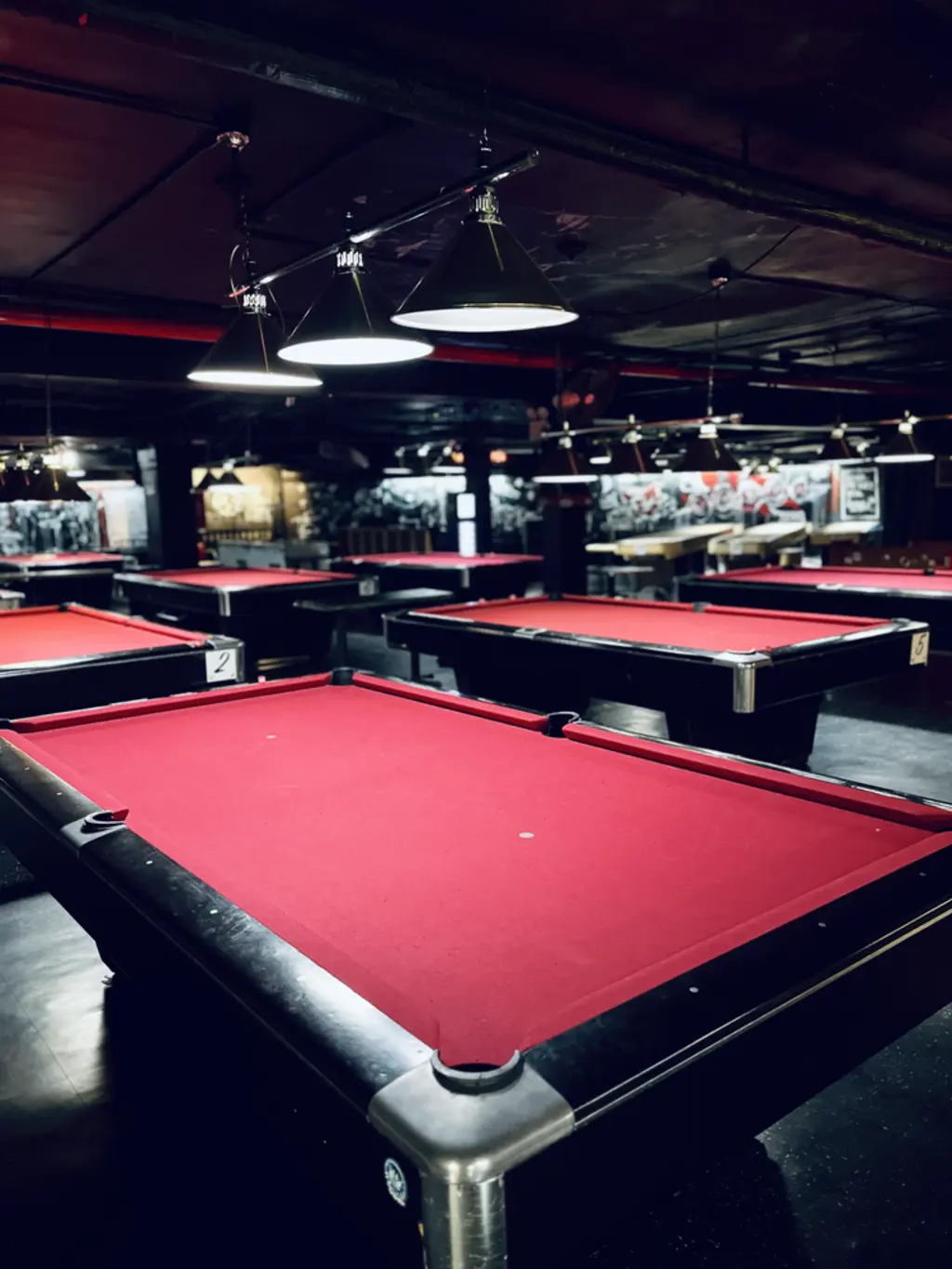 A room with multiple red pool tables and hanging lights, with a graffiti-covered wall in the background.