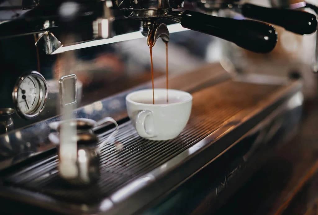 Close-up of espresso pouring from a professional machine into a white cup on a metal tray, with a frothing pitcher nearby, captured in a cozy café setting.