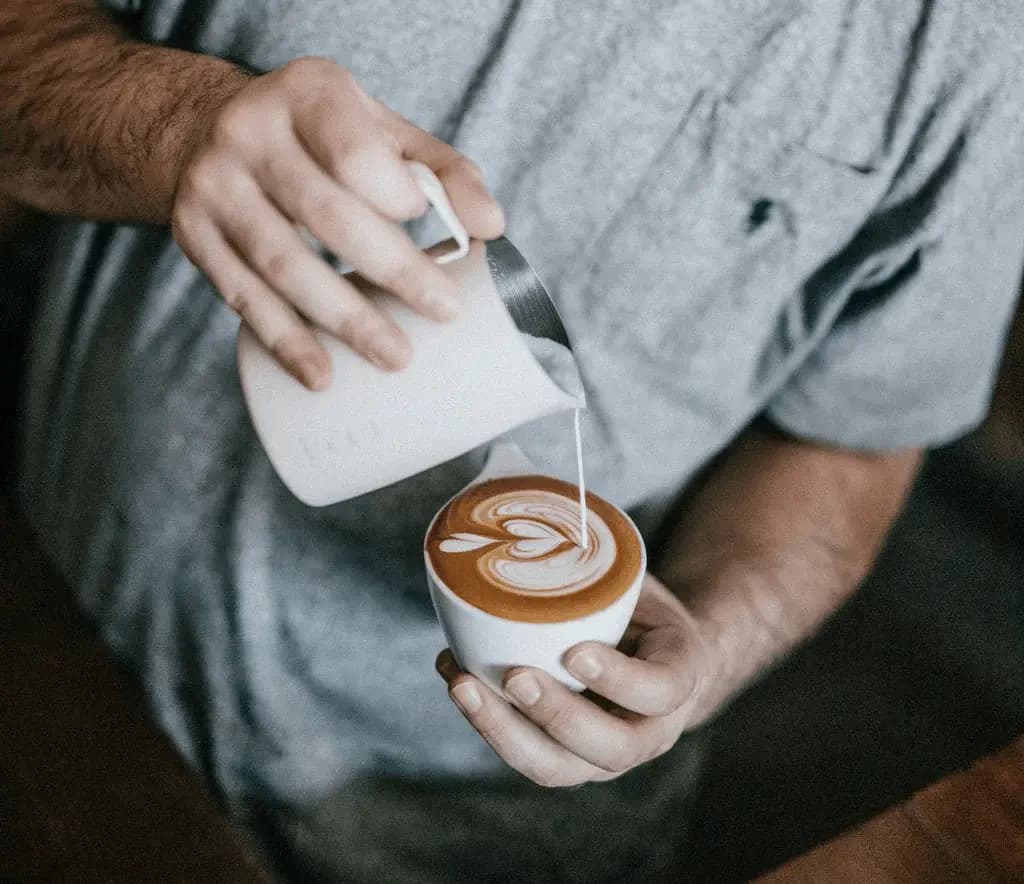 Barista pouring steamed milk into a cup of coffee to create a latte art heart design, showcasing skill and attention to detail in a cozy café atmosphere.