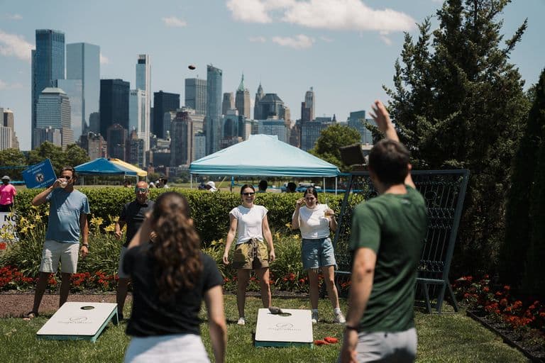 People playing cornhole on a sunny day with the NYC skyline in the background and tents set up in the park.
