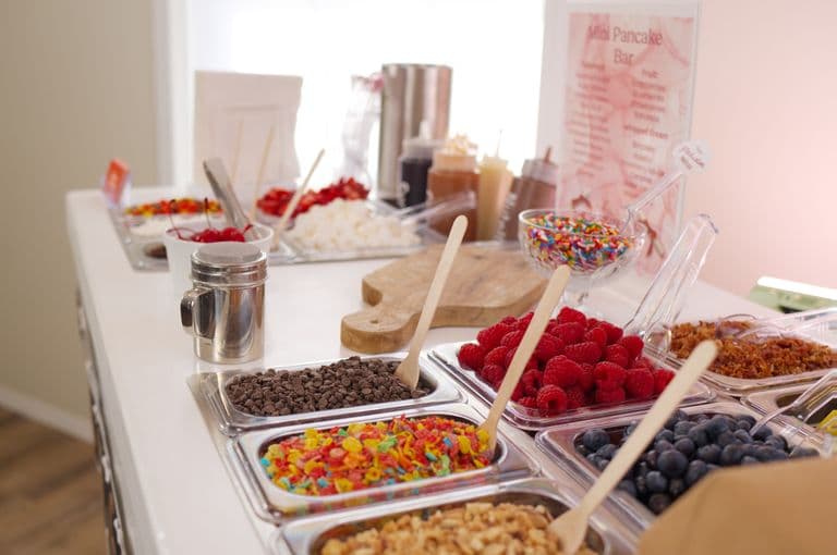 Mini pancake topping bar with fresh berries, sprinkles, chocolate chips, marshmallows, cereal, syrup bottles, and serving spoons laid out on a counter.