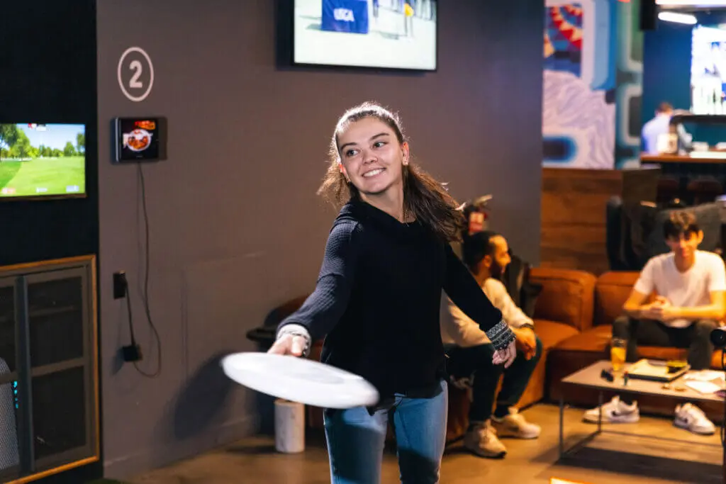 Young woman playing with a frisbee indoors, smiling as she throws it. Others are seated in the background, watching the activity.