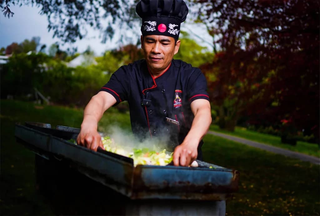 Hibachi chef grilling vegetables outdoors, surrounded by greenery, with steam rising from the hot grill.