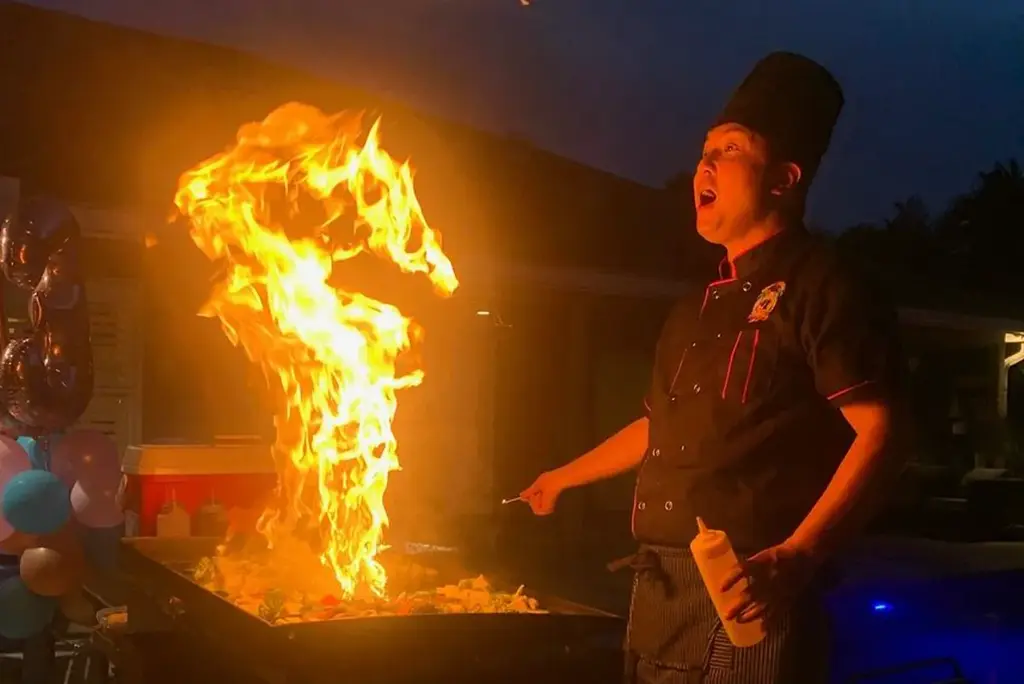 Hibachi chef in action, creating a dramatic fire display on a grill at an evening outdoor event with guests watching.
