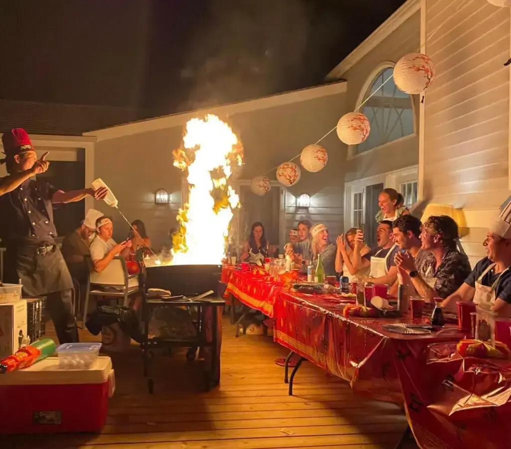 A lively backyard gathering with a hibachi chef cooking on a grill, flames rising high as guests enjoy the show.