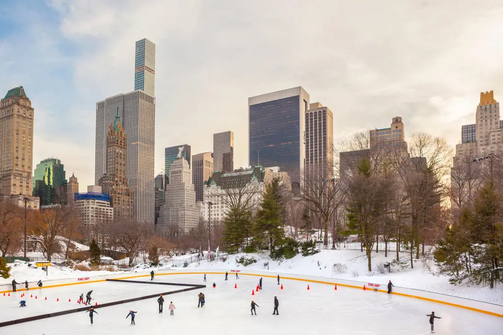 a daytime view of people ice skating at a rink in central park with building in the background
