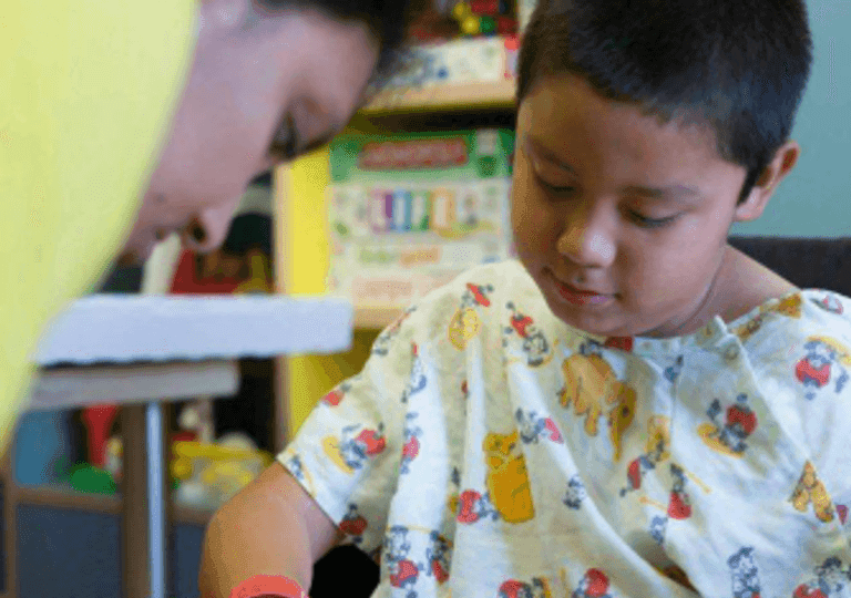 Child in a hospital gown focusing on a craft activity with support from an adult.