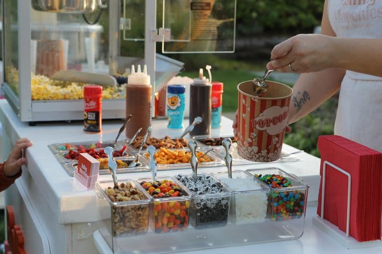 Close-up of a “Rolling Munchies” snack cart offering a popcorn bar with assorted toppings like candy, nuts, pretzels, and sauces, as a person fills a striped popcorn bucket.