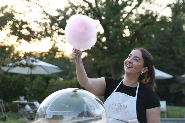 A smiling vendor wearing a “Rolling Munchies” apron spins a large pink cotton candy outdoors, with trees and patio umbrellas softly blurred in the background.