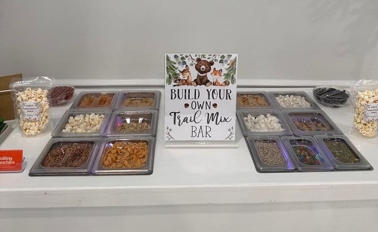 Self-serve trail mix bar setup with assorted snacks and toppings displayed in trays, featuring a “Build Your Own Trail Mix Bar” sign.