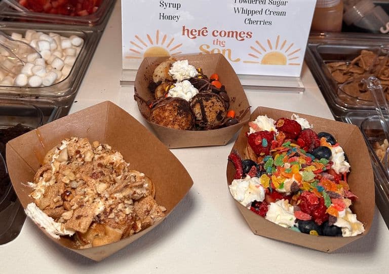 Close-up of a mini pancake bar setup with toppings like fruit, drizzles, and cereals, displayed beside three trays of decorated mini pancakes.