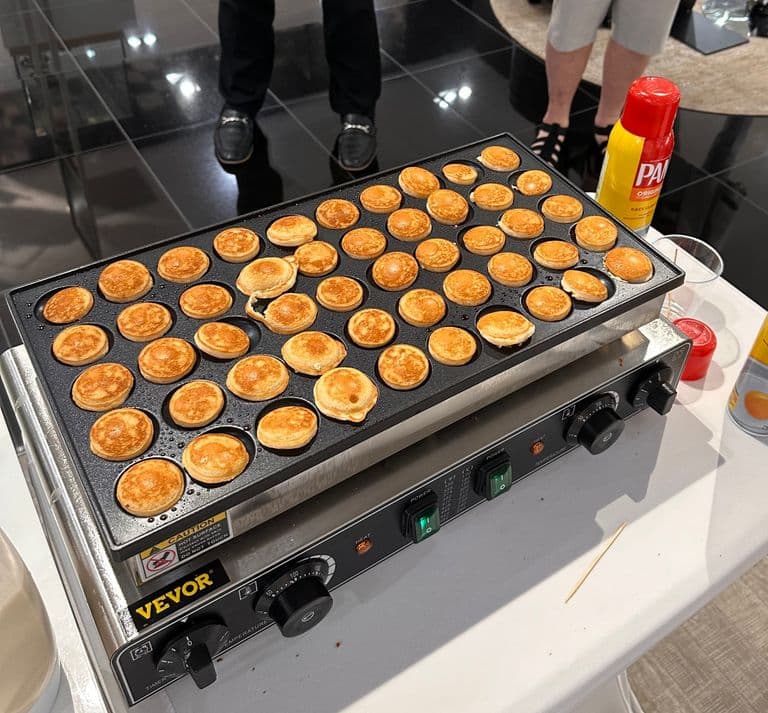 Close-up of a mini pancake bar setup with toppings like fruit, drizzles, and cereals, displayed beside three trays of decorated mini pancakes.