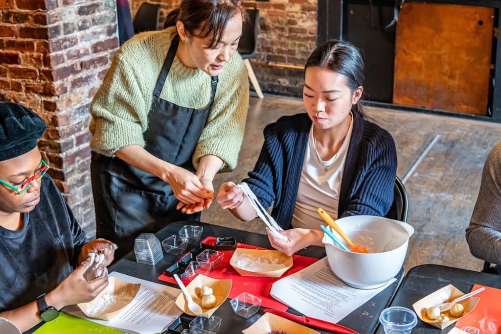Instructor assisting a participant in a Japanese sweets making class, with various ingredients and tools on the table.