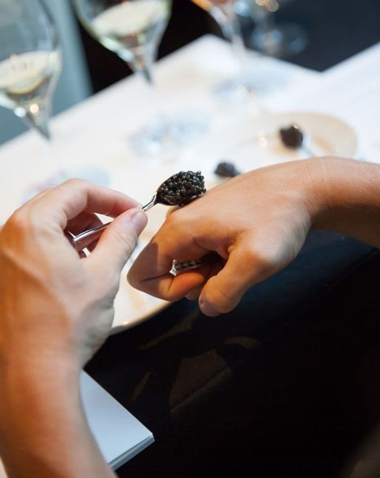 A person holds a spoonful of caviar over their hand during a tasting, with wine glasses and a plate in the background.