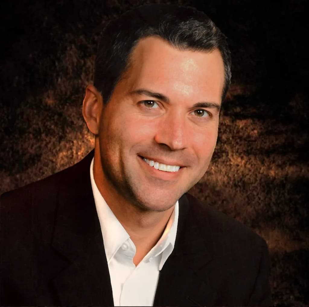 A professional headshot of a man in a dark suit, smiling with a dark, textured background.