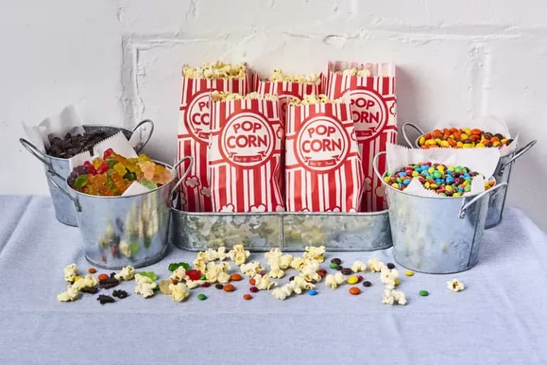 Popcorn bar with red striped bags, candy toppings in metal buckets, and scattered popcorn on a blue tablecloth.