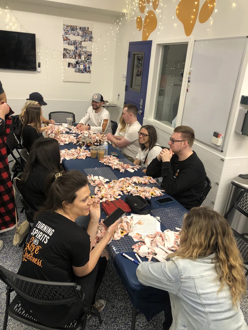 Group of people seated around a table filled with pieces of fleece fabric, smiling and chatting while crafting no-sew blankets together.