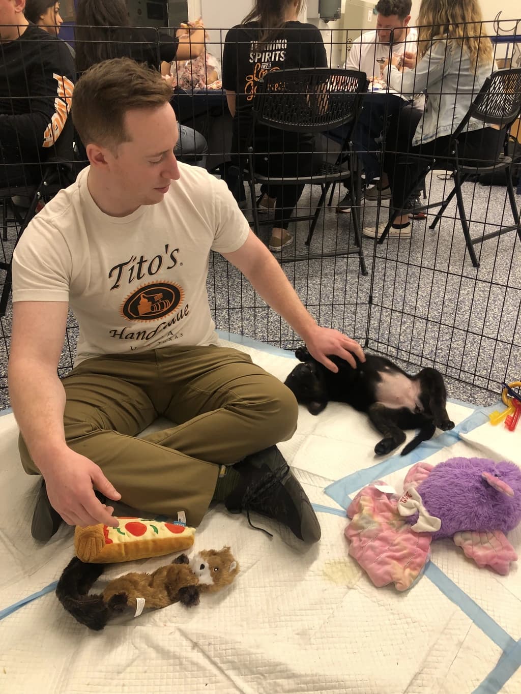 Man in a Tito's Handmade Vodka shirt sits on the floor with a puppy, surrounded by dog toys, smiling and gently petting the puppy.