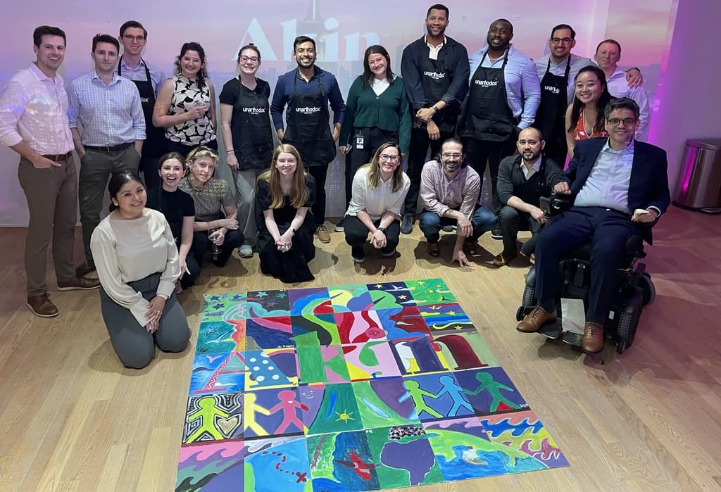 Group of people proudly standing behind a colorful mural they collectively painted, featuring vibrant figures and abstract designs.