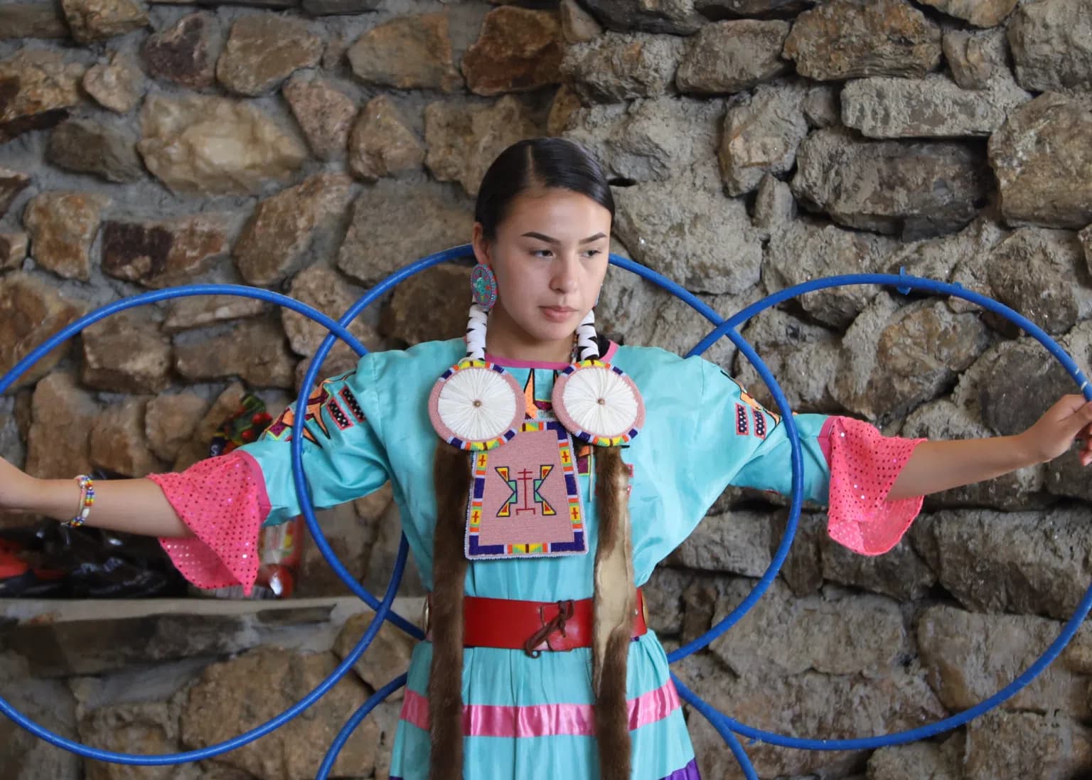 A young Native American woman in traditional regalia performing a hoop dance, with colorful hoops surrounding her.