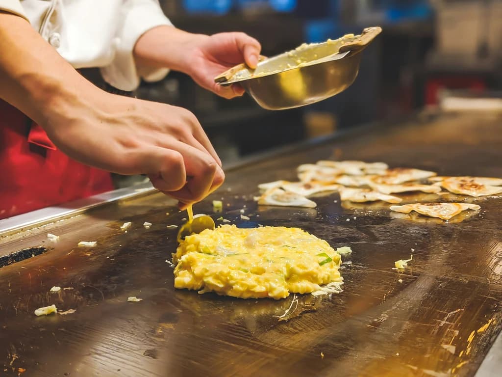 A chef cooking an omelette on a flat-top grill, with other food items cooking nearby.
