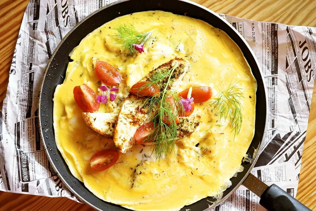 A freshly cooked omelette in a pan, topped with cherry tomatoes, herbs, and edible flowers, placed on a wooden table.