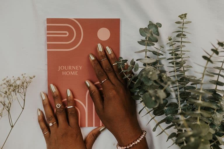Hands with metallic nails and rings resting on a “Journey Home” journal, surrounded by eucalyptus and baby's breath.