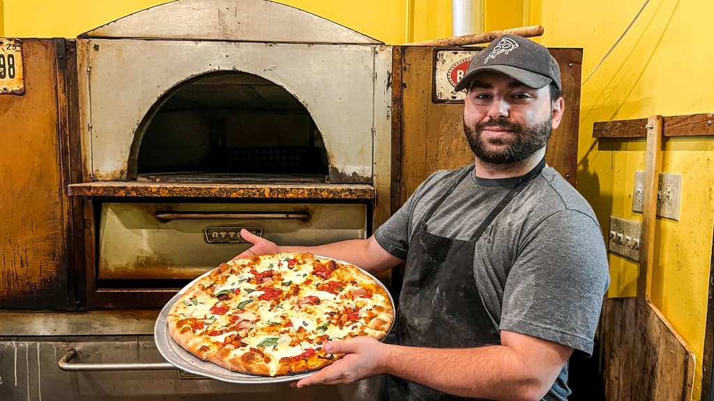 a man in front of a pizza oven, holding a freshly made pizza