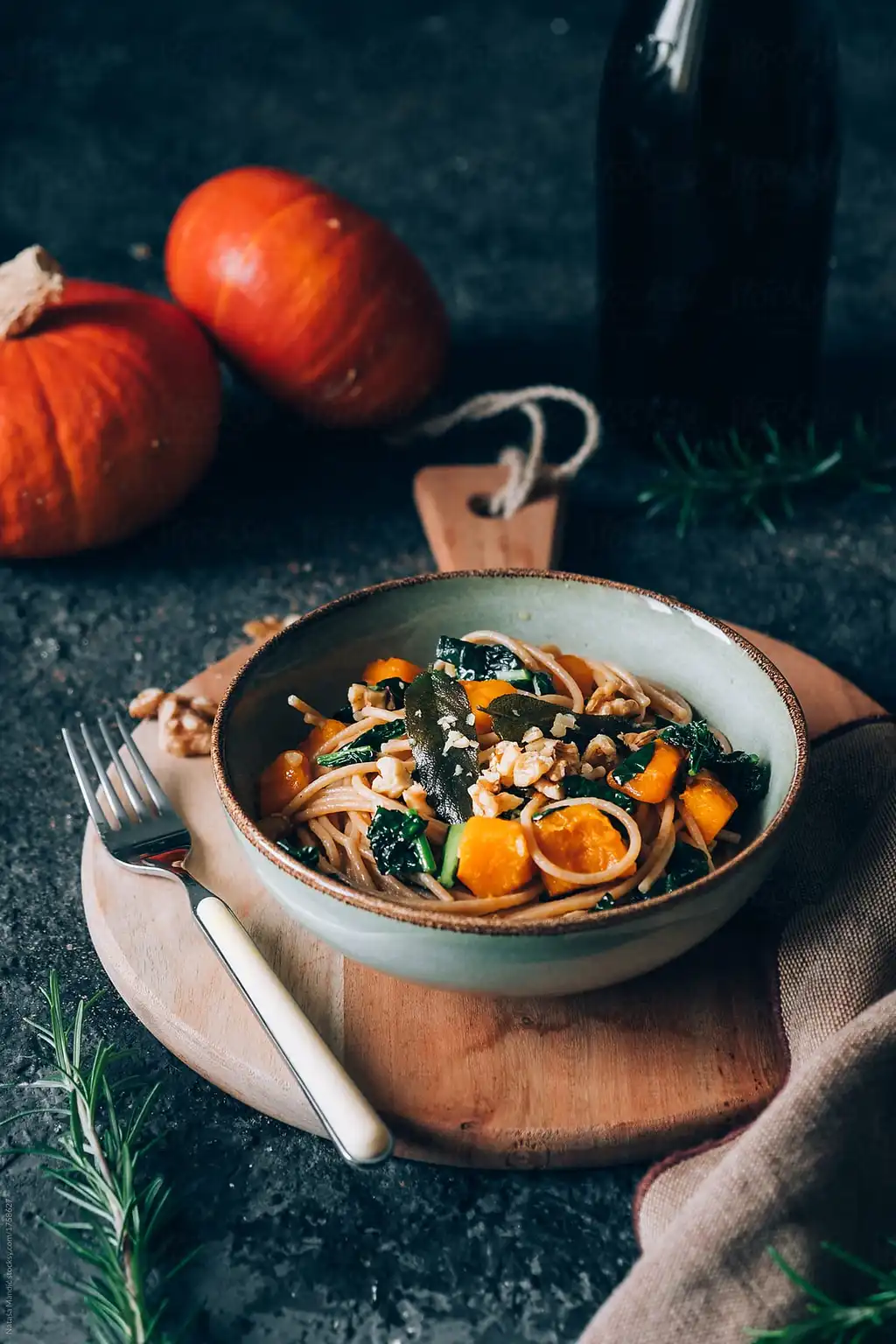 A rustic bowl of pasta with pumpkin chunks, kale, and walnuts, accompanied by two small pumpkins and rosemary on a wooden board.