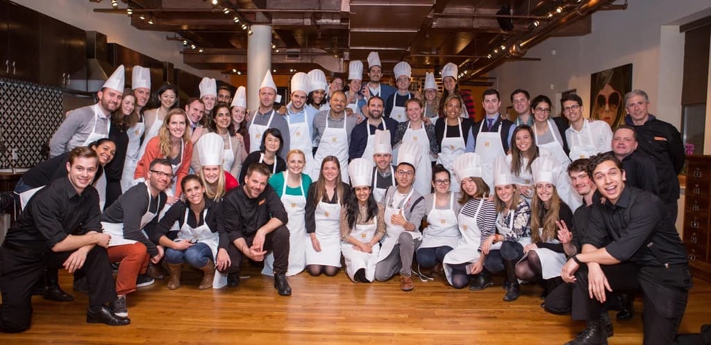 A large group of participants in chef hats and aprons pose together, smiling, in a cooking studio after a group cooking event.