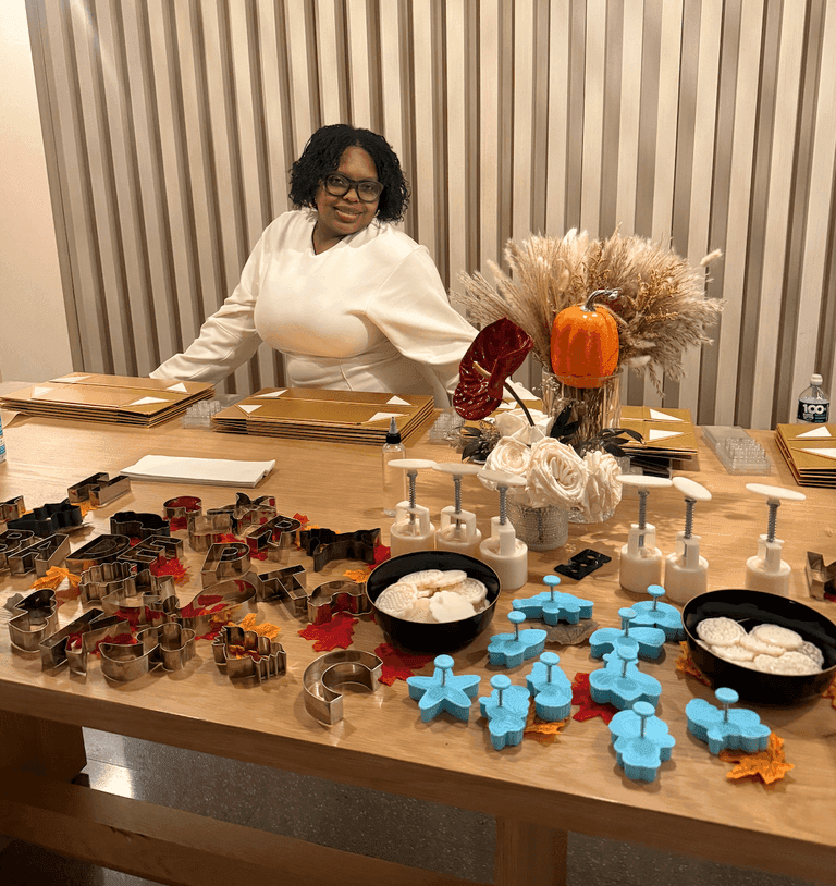 A woman in a white blouse stands behind a table set up for a cookie decorating workshop, featuring cookie cutters, molds, and fall-themed decorations.