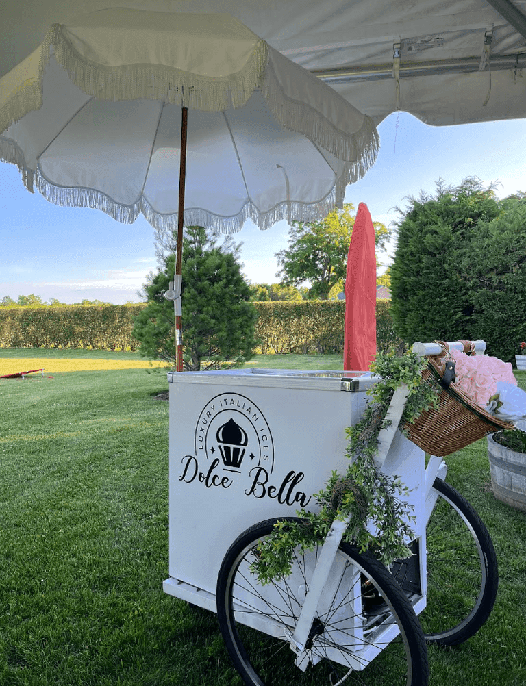 White Dolce Bella Italian ices cart with umbrella on a lawn, decorated with flowers and a basket.