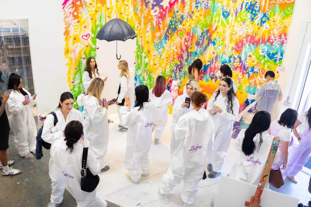 A group of people in white protective suits mingling in a brightly decorated room with a multicolored wall mural, some taking photos and chatting.