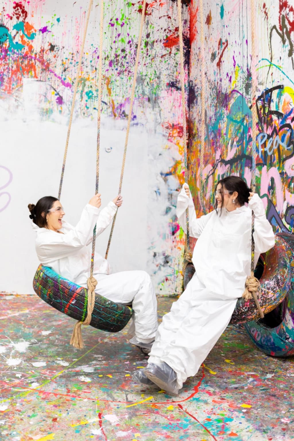 Two women in white protective suits joyfully swinging on colorful, paint-splattered tire swings in a vibrant, graffiti-covered room.