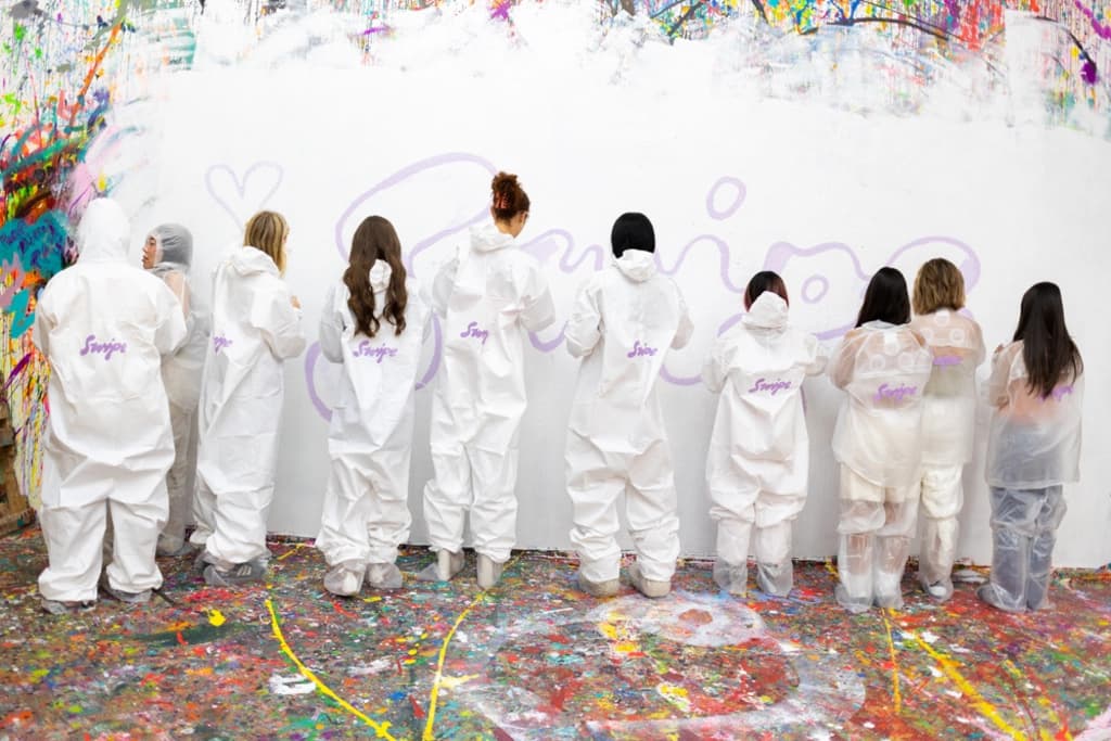 A line of people wearing white protective suits, facing a wall, creatively painting and adding to a vibrant, abstract mural in an art-filled space.