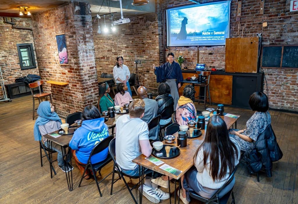 Group of people attending a tea ceremony presentation in a rustic room with brick walls, learning about the art of Matcha and Samurai.
