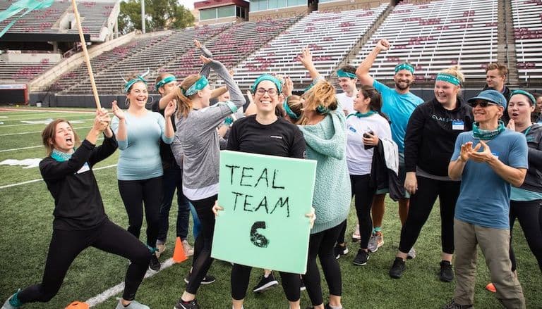 Team wearing teal bandanas celebrates on a stadium field, holding a large “Teal Team 6” sign and cheering enthusiastically.