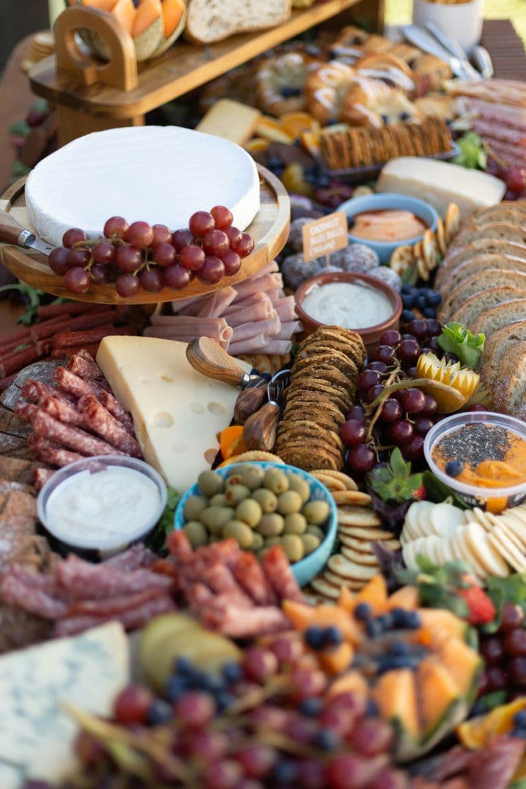 Close-up of an abundant grazing table featuring a large wheel of brie, assorted cheeses, cured meats, fresh fruits, olives, dips, crackers, and artisan bread.