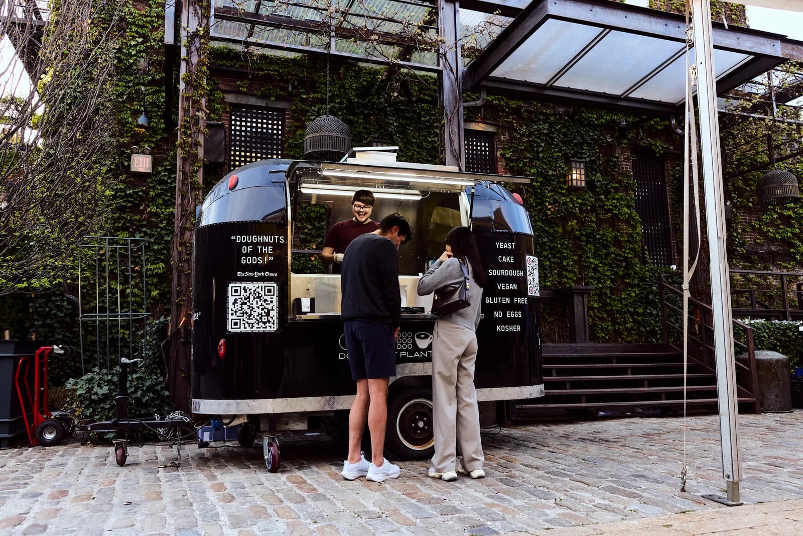 Black Doughnut Plant Airstream trailer serving guests through an open window beside an ivy-covered wall