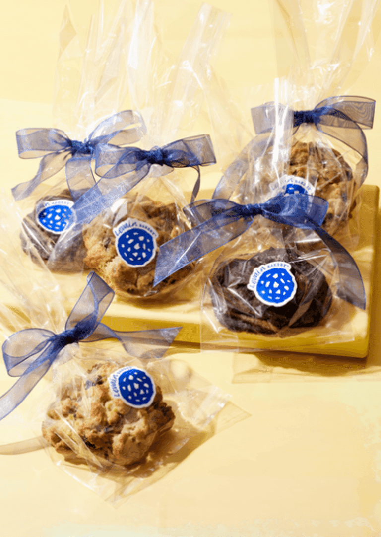 Levain Bakery cookies wrapped in clear bags with blue ribbons and branded stickers, displayed on a yellow plate against a bright background.