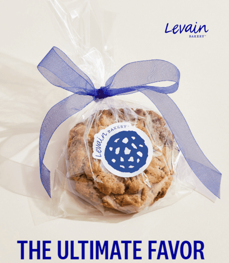 A Levain Bakery cookie wrapped in clear plastic with a blue ribbon and branded sticker, with "THE ULTIMATE FAVOR" text on a white background.