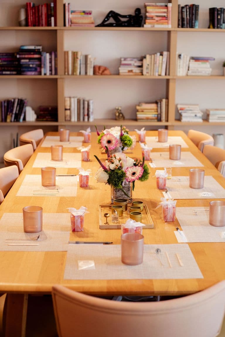 A beautifully set table for a workshop, featuring floral centerpieces, gift bags, candles, and neatly arranged materials in a cozy book-filled space.